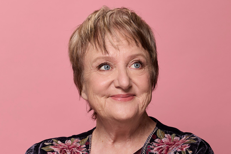 Studio portrait of Australian comedian and actress Denise Scott smiling against a solid pink background, wearing an embroidered floral dark top.